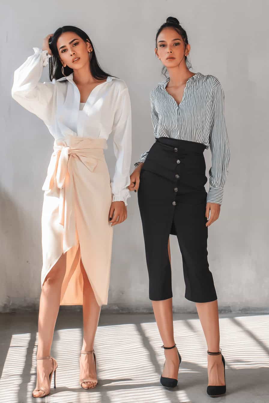 Zwei Frauen in eleganten Blusen und Röcken, stehend in lichtdurchfluteter Umgebung.