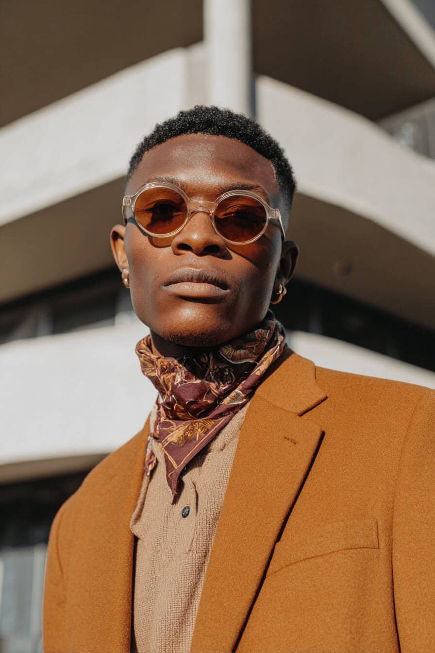 Mann mit braunem Blazer, Sonnenbrille und gemustertem Halstuch vor moderner Architektur