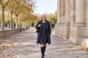 Frau in marineblauem Etuikleid, langem marineblauem Wollmantel und Stiefeln vor einem Steingebäude im Herbst