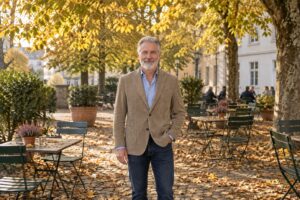 Mann in camelfarbenem Cordsakko, hellblauem Hemd und dunkler Jeans auf einer Caféterrasse im Herbst