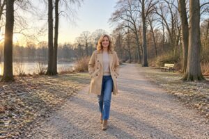 Frau in camelfarbenem Trenchcoat, sandfarbenem Feinstrickpullover und Jeans auf einem Parkweg im Winter