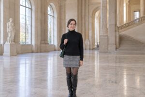 Frau in schwarz-weiß kariertem Minirock, schwarzem Rollkragenpullover und Overknee-Stiefeln in einer hellen Museumshalle