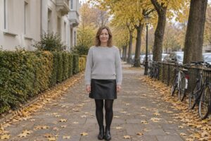 Frau in schwarzem Lederrock, blickdichter Strumpfhose, Stiefeletten und hellgrauem Grobstrickpullover auf einem herbstlichen Gehweg